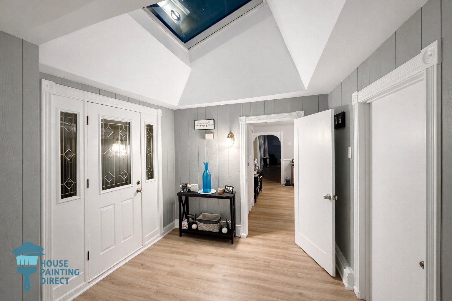 A bright home entryway featuring gray paneled walls, white decorative trim, and light wood flooring. The space is illuminated by a large overhead skylight set into a white vaulted ceiling, with a "House Painting Direct" logo in the bottom left corner.
